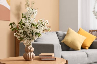 Oturma odasındaki kanepenin yanındaki sehpanın üzerine kitap konmuş beyaz çiçek açan bir vazo.