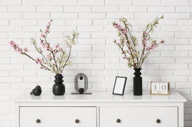 Farklı dekoru olan bir giysi ve oturma odasındaki beyaz tuğla duvarın yanında çiçek açan bir vazo.