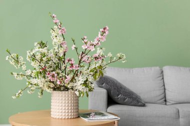 Oturma odasındaki yeşil duvara yaslanmış sehpanın yanında dergi bulunan çiçek açan bir vazo.