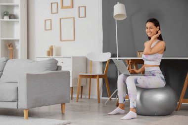 Evde spor yaparken dizüstü bilgisayar kullanan genç bir kadın.