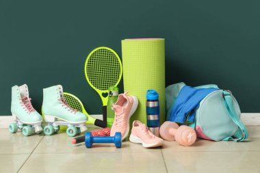 Farklı spor aletleri, patenler ve mavi duvarın yanındaki döşemede spor ayakkabıları.