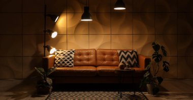 Kahverengi kanepesi, masası, çiçekleri ve parlayan lambaları olan karanlık oturma odasının içi.