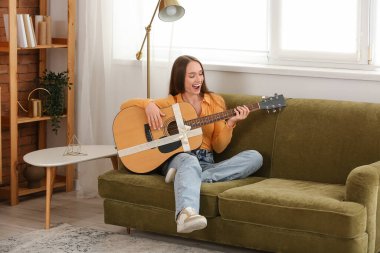 Happy young woman with gift guitar sitting on sofa at home