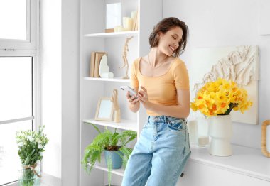 Young woman using mobile phone with daffodils in vase on commode at home