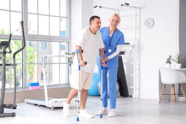 Kadın fizyoterapist ve rehabilitasyon merkezinde yürüyen koltuk değnekleriyle genç bir adam.