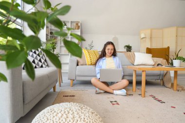 Genç Asyalı kadın evdeki gri kanepenin yanındaki halıda dizüstü bilgisayar kullanıyor.