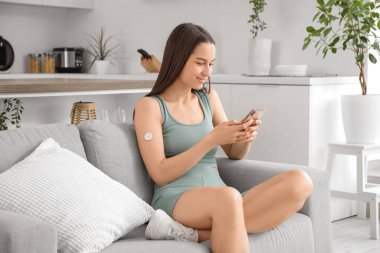 Glikoz sensörü olan şeker hastası genç bir kadın evdeki koltukta cep telefonu kullanıyor.