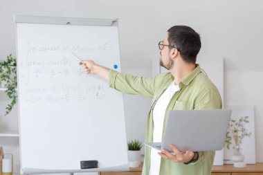 Dizüstü bilgisayarı ve formülleri olan genç erkek matematik öğretmeni evde online ders veriyor.