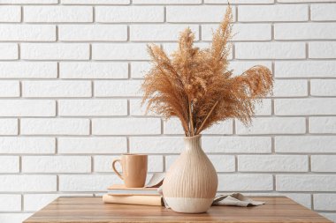 Kuru kamışlı vazo, kitaplar ve fincanlar açık tuğla duvara karşı ahşap masada.