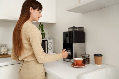 Güzel, genç, mutlu bir kadın mutfakta makineyle kahve yapıyor.