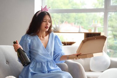 Evdeki doğum günü partisinden sonra elinde şişe ve pizza kutusuyla kanepede oturan taç giymiş genç Asyalı kadın.