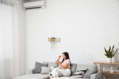 Oturma odasındaki beyaz tuğla duvarın yanındaki koltukta klimalı uzaktan kumandalı genç bir kadın.