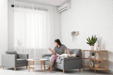 Klimalı uzaktan kumandalı genç bir kadın evdeki beyaz tuğla duvarın yanındaki kanepede oturuyor.