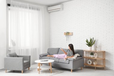 Klimalı uzaktan kumandalı genç bir kadın evdeki beyaz tuğla duvarın yanındaki kanepede yatıyor.