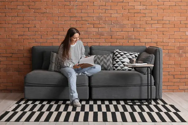 Genç ve güzel bir kadın evdeki siyah koltukta dergi okuyor.