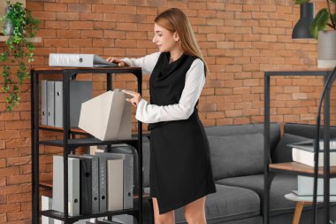 Genç iş kadını, ofisteki raftan evrak klasörünü alıyor.