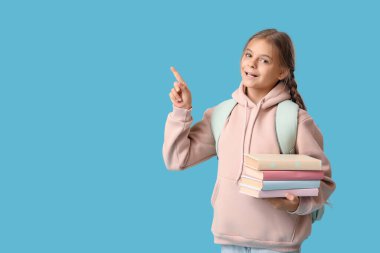 Kitapları ve sırt çantasıyla mavi arka plandaki bir şeyi gösteren tatlı bir kız öğrenci.