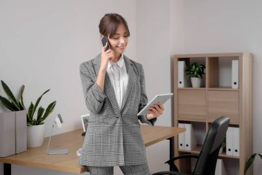Güzel genç iş kadını cep telefonuyla konuşuyor ve modern tablet bilgisayarını ofisinde tutuyor.