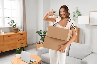 Kulaklıklı, evdeki paketleri gösteren güzel, genç ve mutlu bir kadın.