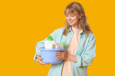 Sarı arka planda çamaşır deterjanı şişeleriyle dolu bir leğen tutan genç kadın.