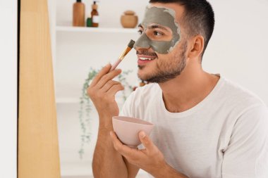 Yakışıklı genç adam banyoda aynanın yanında kozmetik kilden maske takıyor.