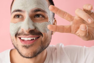 Yüzünde kozmetik kil maskeli yakışıklı genç adam pembe arka plana karşı zafer jesti gösteriyor.