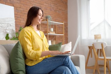 Evdeki gri koltukta dizüstü bilgisayar kullanan genç bir kadın.