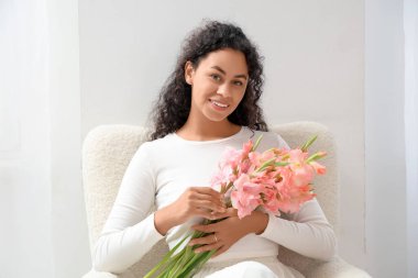 Güzel, mutlu, Afro-Amerikan bir kadın. Elinde pembe gladyatör çiçekleri var. Evdeki koltukta oturuyor.