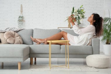 Güzel, mutlu, Afro-Amerikan bir kadın. Elinde bir buket beyaz zambak çiçeğiyle evdeki koltukta oturuyor.