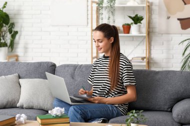 Evdeki kanepede oturan dizüstü bilgisayarı ve defteri olan güzel mutlu kadın yazar.