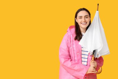 Yağmurluk giymiş, sarı arka planda şemsiyesi olan mutlu genç bir kadın.