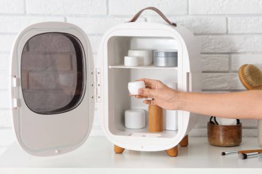 Beyaz tuğla duvarın yanındaki masada krema kavanozu ve küçük kozmetik buzdolabı olan bir kadın eli. Yakın çekim.