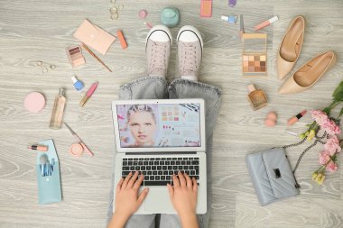 Farklı kozmetik ürünleri ve aksesuarları olan genç bir kadın açık ahşap zeminden online alışveriş için dizüstü bilgisayar kullanıyor.