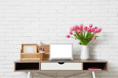 Modern dizüstü bilgisayarlı şık çalışma alanı, bir fincan kahve ve beyaz tuğla duvarın yanında güzel lale çiçekleri.