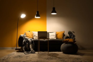 Karanlık oturma odasının içinde kanepe, dizüstü bilgisayar ve parlayan lambalar var.