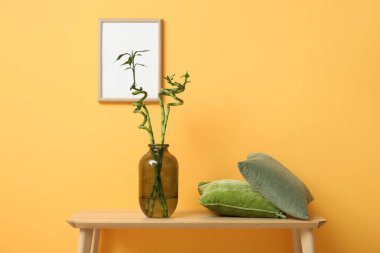 Turuncu duvarın yanındaki tahta bankta bambu dalları ve yastıkları olan vazolar.