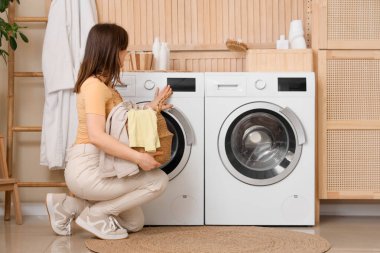 Çamaşır odasında çamaşır makinesinin yanında sepet tutan genç bir kadın.