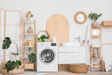 Modern çamaşır makinesi, hasır sepeti, raf ünitesi ve banyo aksesuarları olan şık bir çamaşırhane.