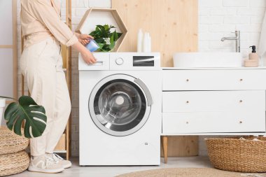 Çamaşır odasındaki çamaşır makinesine çamaşır deterjanı koyan genç bir kadın.