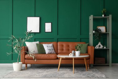 Oturma odasındaki yeşil duvarın yanında masası ve rafı olan kahverengi deri kanepe.