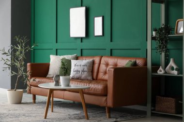 Oturma odasındaki yeşil duvarın yanında masası ve rafı olan kahverengi deri kanepe.