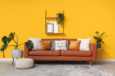 Yumuşak yastıklı kahverengi deri kanepe ve oturma odasındaki sarı duvarın yanında puf.