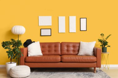 Yumuşak yastıklı kahverengi deri kanepe ve oturma odasındaki sarı duvarın yanında puf.