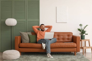 Modern dizüstü bilgisayarlı genç adam oturma odasının yanındaki kahverengi deri koltukta oturuyor.