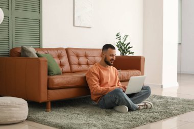 Modern dizüstü bilgisayarlı genç adam oturma odasındaki kahverengi deri koltuğun yanında oturuyor.