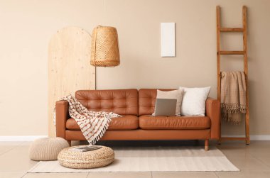 Modern dizüstü bilgisayarı olan kahverengi deri kanepe ve oturma odasındaki bej duvarın yanında yumuşak puflar.