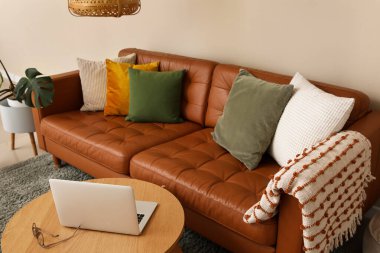 Kahverengi deri kanepe ve oturma odasındaki bej duvarın yanında modern dizüstü bilgisayarı olan ahşap masa.