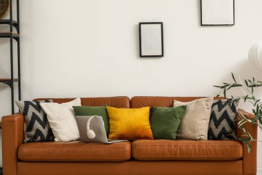 Yumuşak yastıklı kahverengi deri kanepe ve oturma odasındaki beyaz duvarın yanında modern laptop.