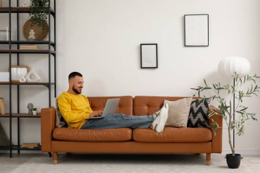 Modern dizüstü bilgisayarlı genç adam oturma odasında kahverengi deri koltukta oturuyor.