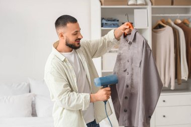 Genç adam yatak odasında buharlı gömlek giyiyor.